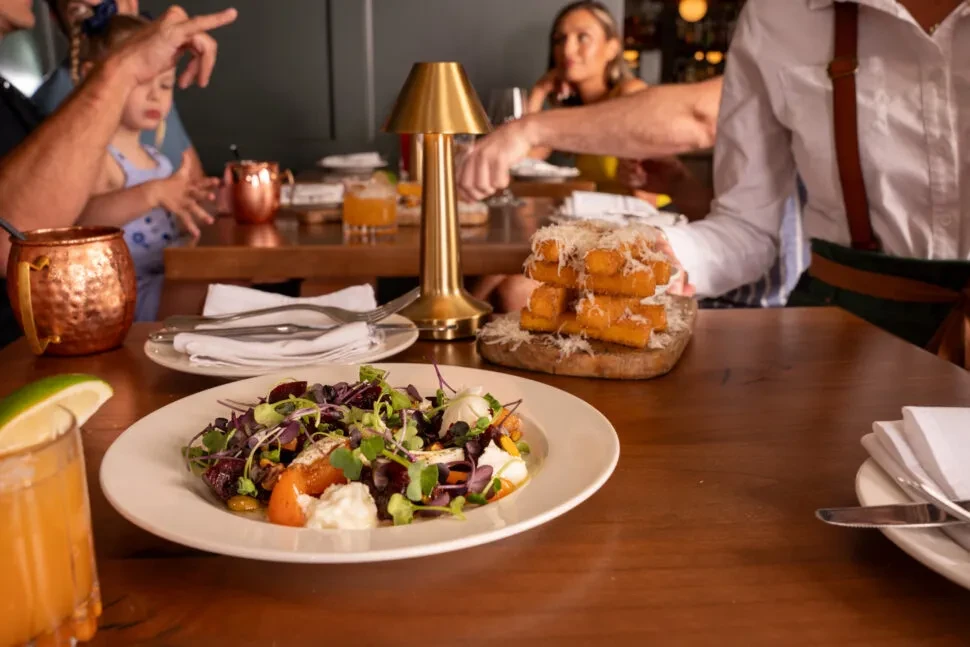 Restaurant table with cocktail, salad, cheesy fries, and blurred diners.
