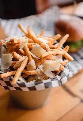 Piled golden fries with cheese shavings in cup, blurred burger behind.