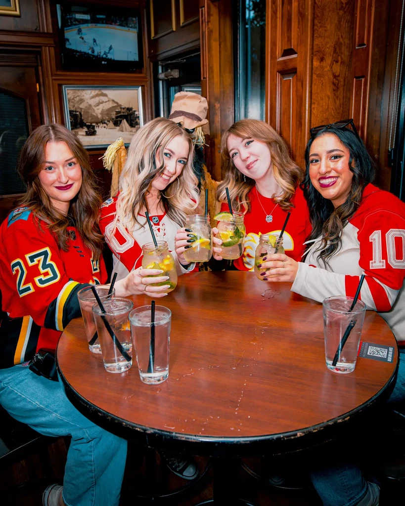 Four young women in hockey jerseys smile, drinking cocktails in a pub.
