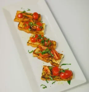 Five bruschetta with grilled bread, tomatoes, and herbs on a white plate.