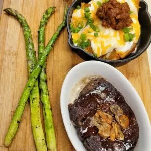 Seared steak, grilled asparagus, and loaded potato skillet on wooden board.
