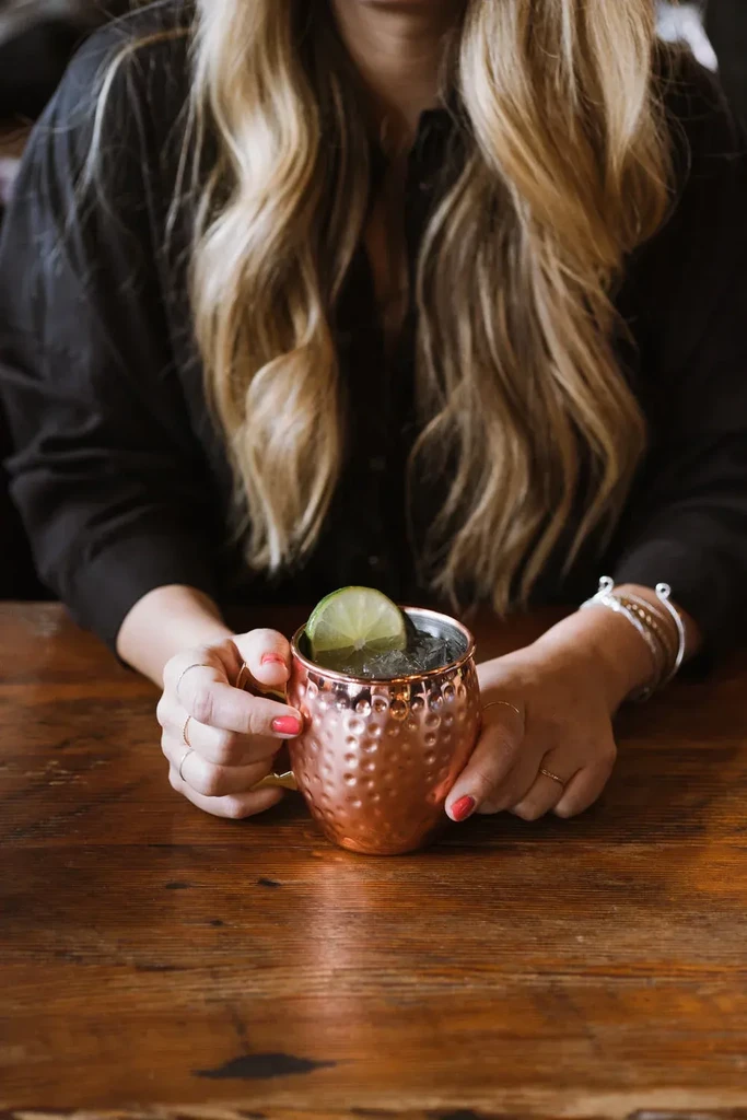 Hands hold hammered copper mug with lime and ice on wood.