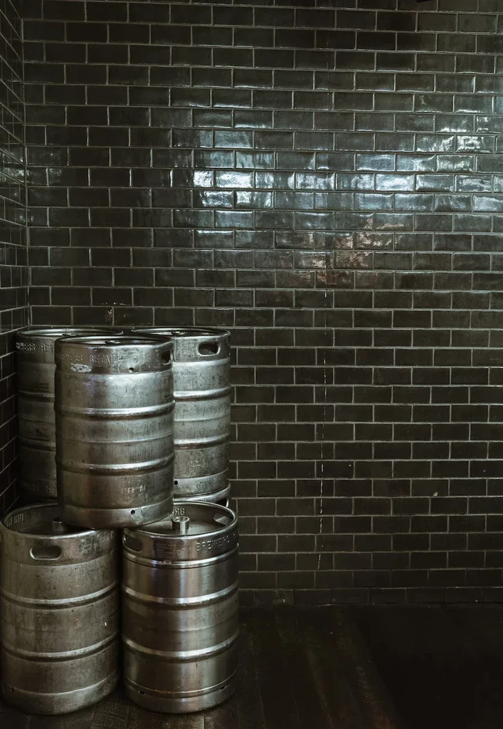 Five worn metallic beer kegs stacked against a dark tile wall.