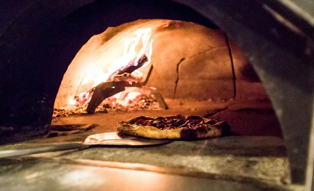Pizza cooking inside a rustic wood-fired oven with active flames.