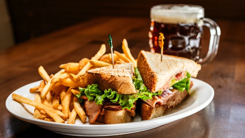 Toasted club sandwich, golden fries, and beer on a wooden table.