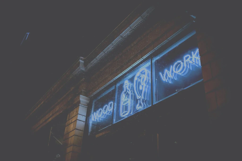 Blue neon signs: WOOD, bottle, WORK, on dark building exterior.