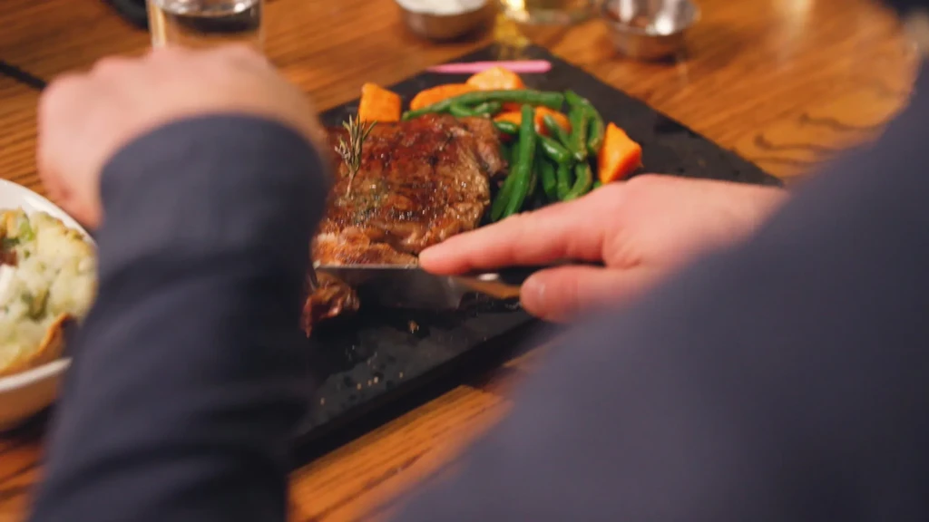Hands cutting a rosemary-garnished steak, green beans, and carrots.