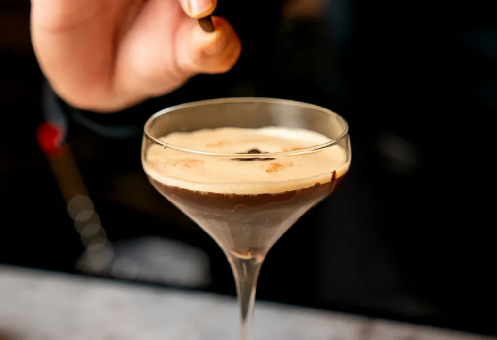 Frothy brown cocktail in martini glass, hand adds coffee bean.