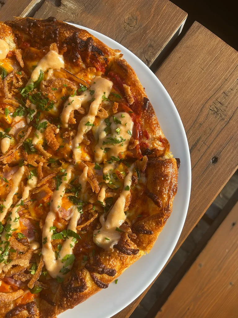 Overhead pizza with ham, crispy onions, herbs, drizzled sauce on wood.