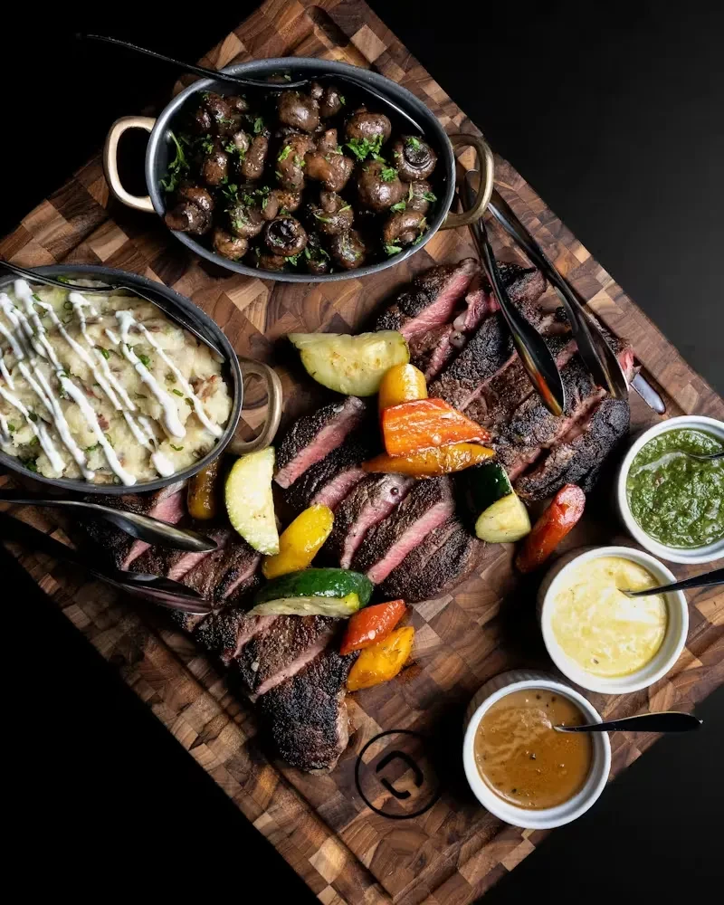 Sliced steak, roasted vegetables, mashed potatoes, mushrooms, sauces on a board.