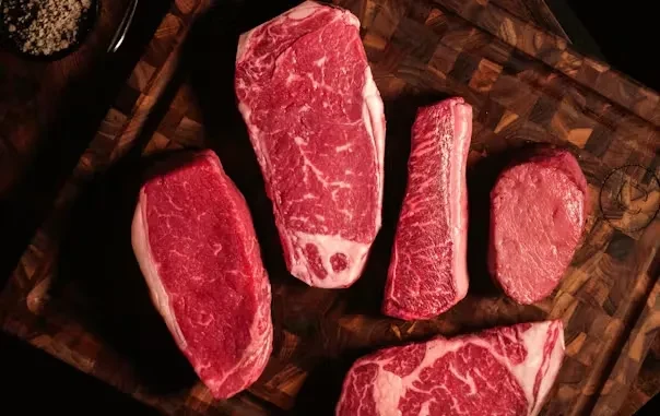 Five raw beef steaks on dark wooden board with seasoning bowl.