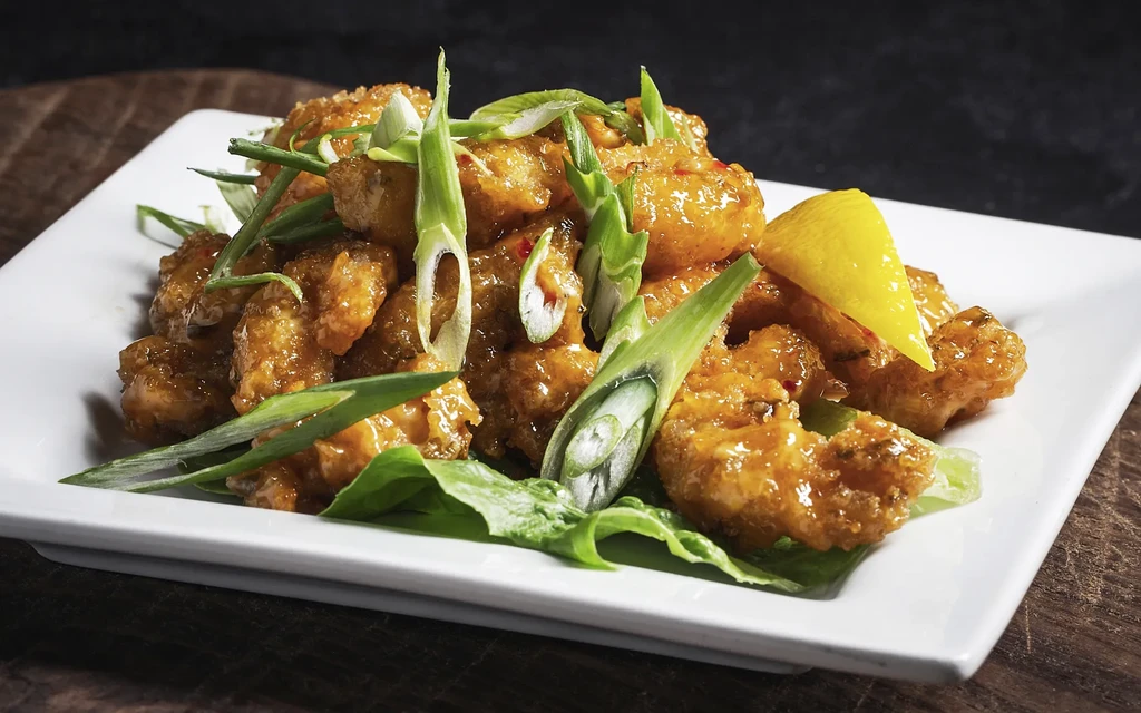 Crispy shrimp in orange sauce with scallions and lemon on a white plate.