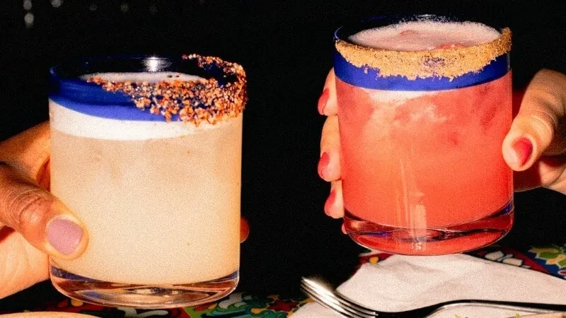 Two hands with colored nails hold frothy and pink rimmed cocktails.