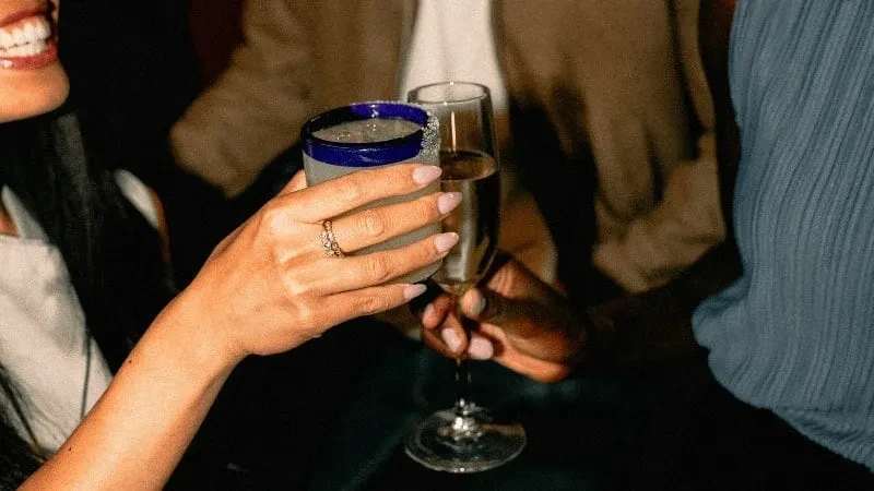 Two hands clinking a blue-rimmed margarita and a champagne flute.