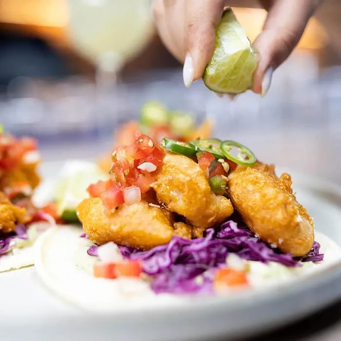 Close-up of a hand squeezing lime over a crispy fish taco with salsa.