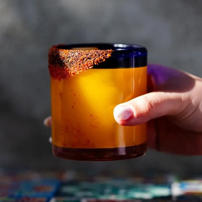 Hand holding orange drink in a blue-rimmed glass with red seasoning.
