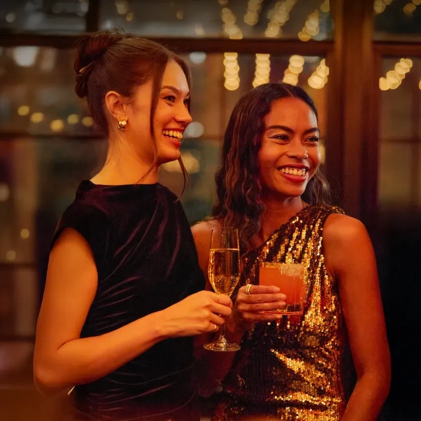 Two smiling women in elegant dresses holding cocktails and champagne.