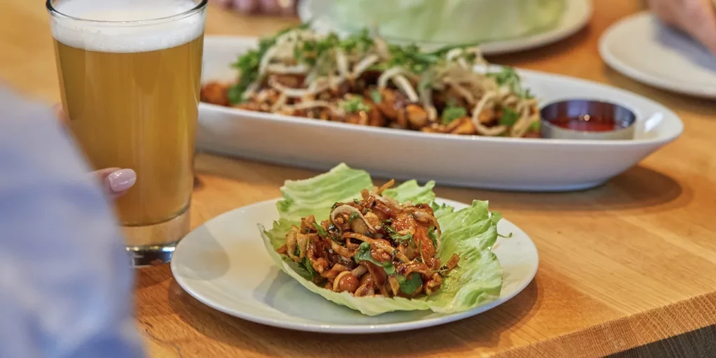 Hand holding beer and a lettuce wrap on a wooden table.