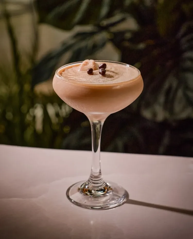 Creamy cocktail in coupe glass with lychee and coffee bean garnish.