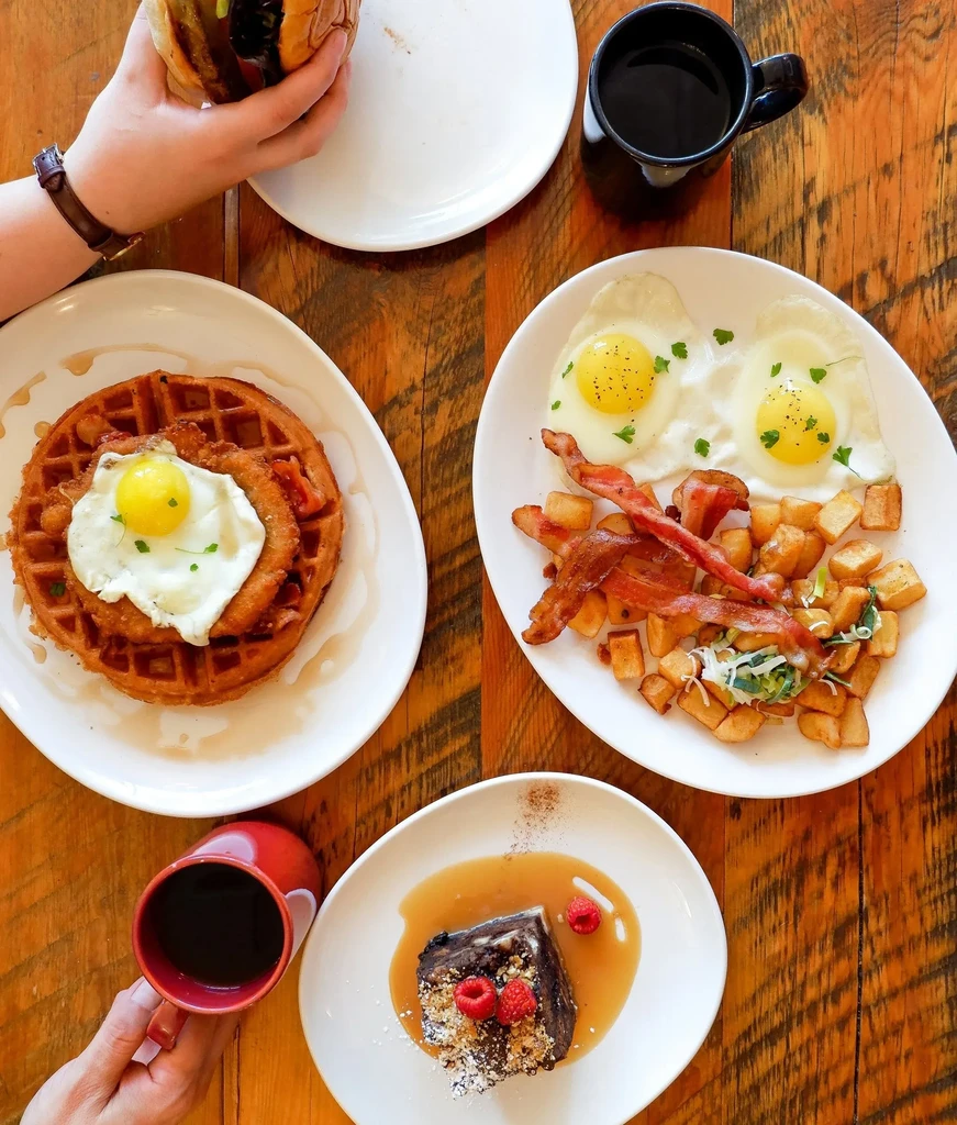 Overhead shot: hands holding food and coffee on a rustic brunch table.