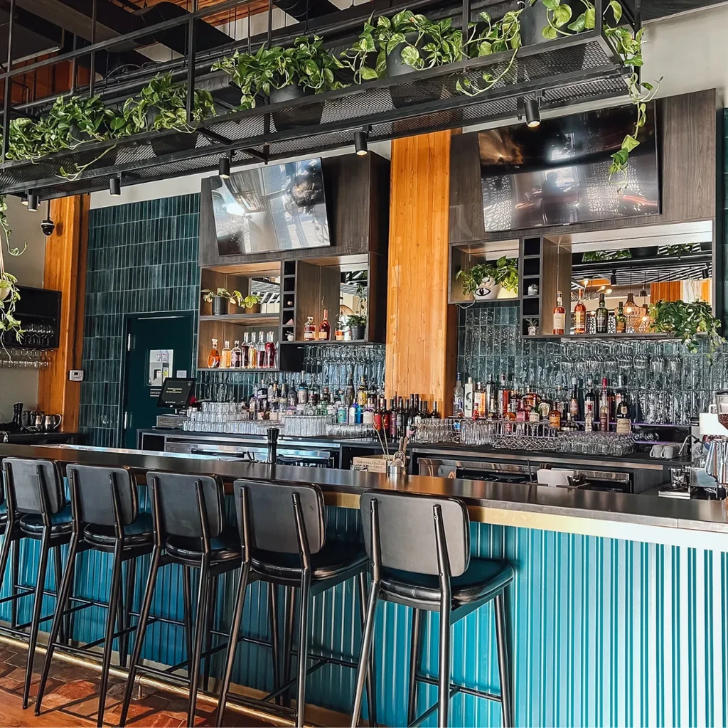 Modern bar with teal fluted counter, dark stools, liquor bottles, and plants.