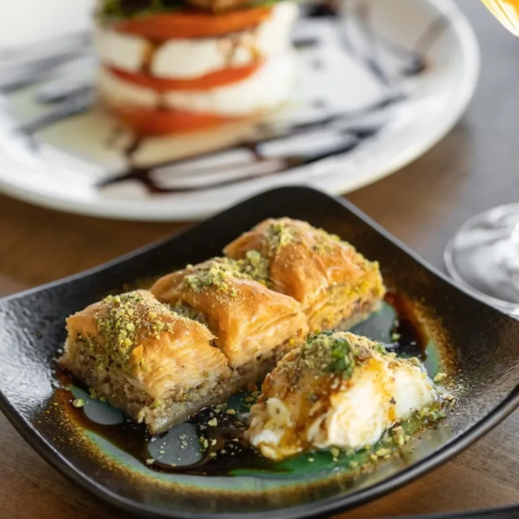 Four syrupy baklava with pistachios and cream; blurred Caprese salad behind.