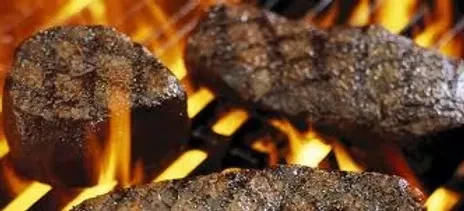Three seared steaks grilling on a grate with intense flames.