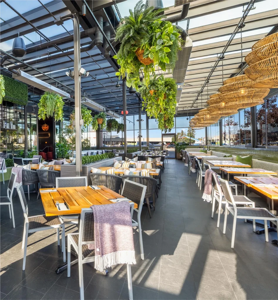 Modern restaurant with greenhouse roof, lush plants, and cozy seating.