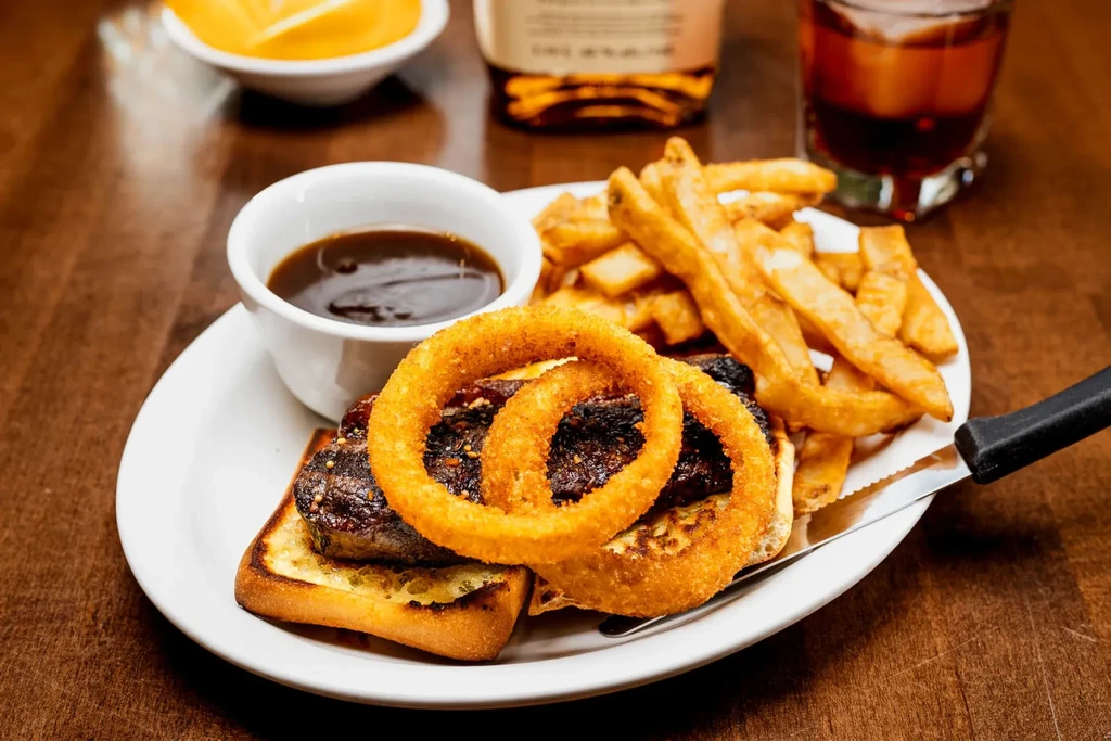 Grilled meat patty on toast, onion rings, french fries, and sauce.