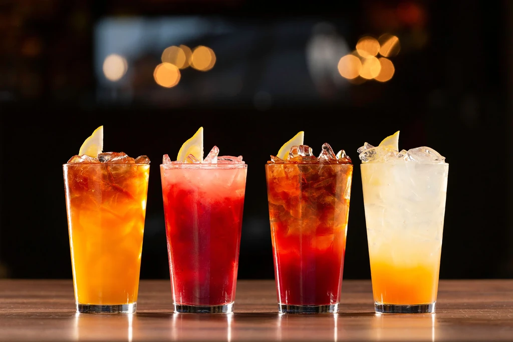 Four colorful iced drinks with lemon wedges on a wooden bar.