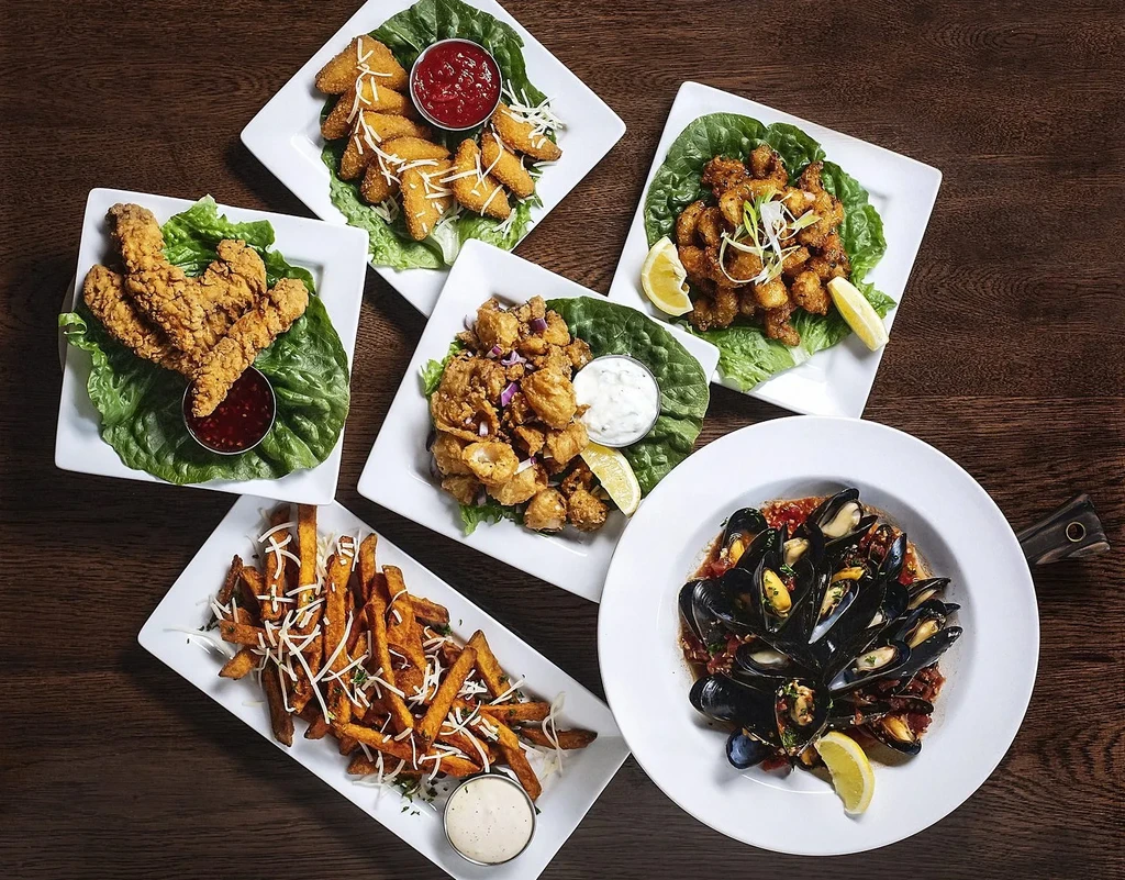 Six distinct fried food dishes and mussels on a dark wooden table.