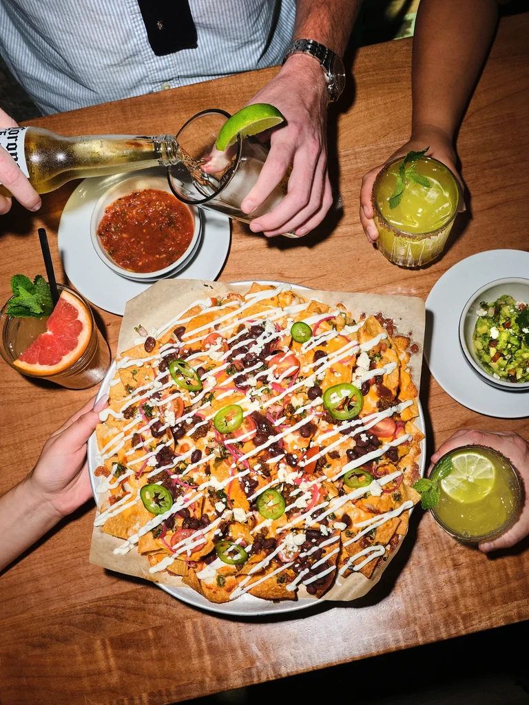 Overhead view of nachos, cocktails, and hands on a wooden table.