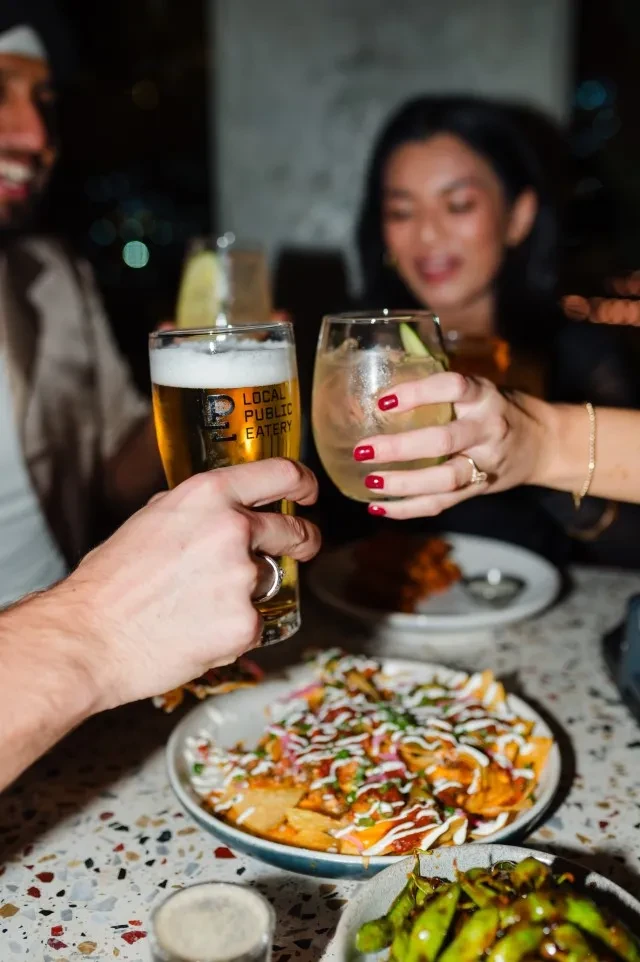 Two blurred people toast beer and cocktail with nachos at a restaurant.