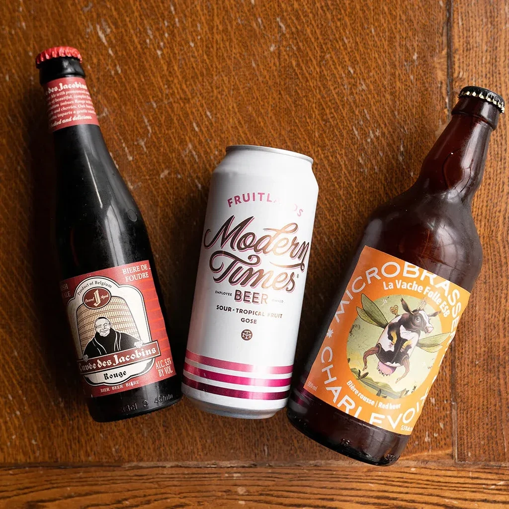 Two beer bottles and a sour beer can rest on wood.