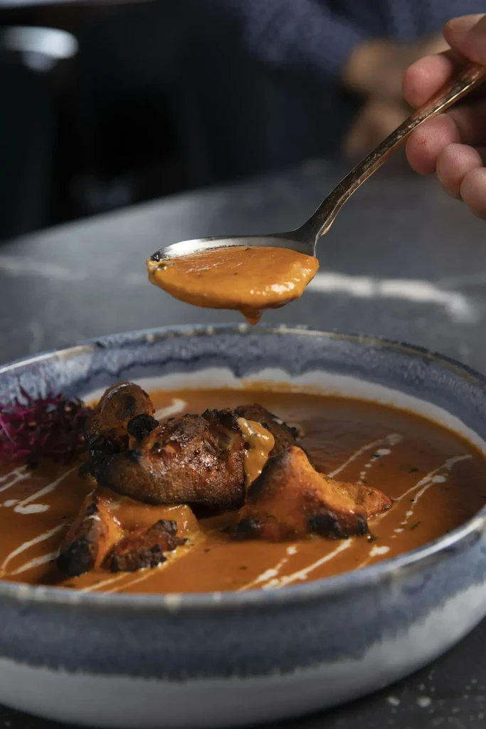 Hand scooping reddish-orange curry with charred protein from patterned bowl.