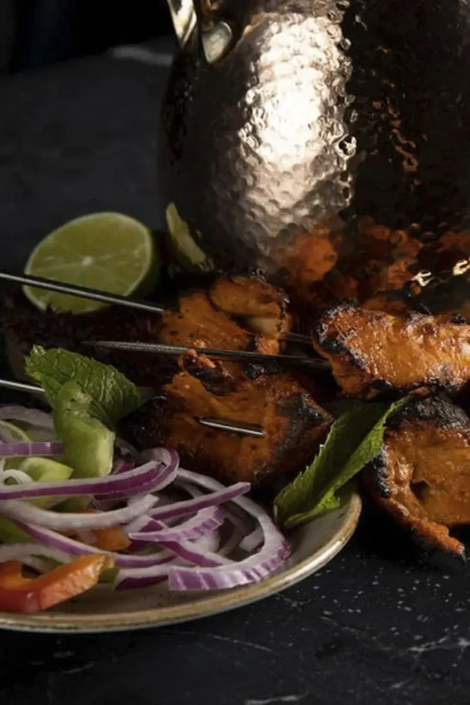 Grilled reddish-orange chicken skewers with onions, mint, lime, and a copper pitcher.