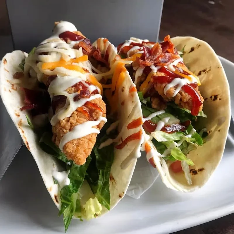 Two loaded fried chicken tacos with cheese, bacon, lettuce, and sauces.