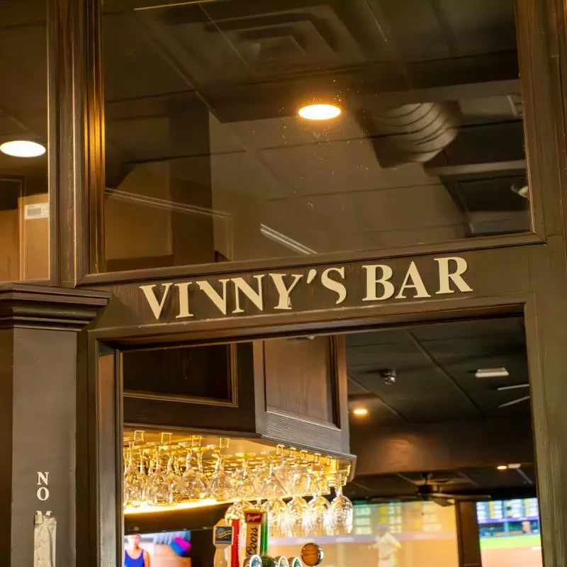 VINNY'S BAR sign, hanging glasses, beer taps, and a TV with baseball.