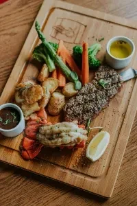 Grilled steak and lobster, roasted vegetables, sauces on wooden board.