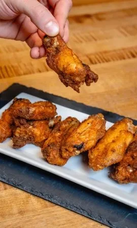 Hand holds golden-brown fried chicken wing from white plate.