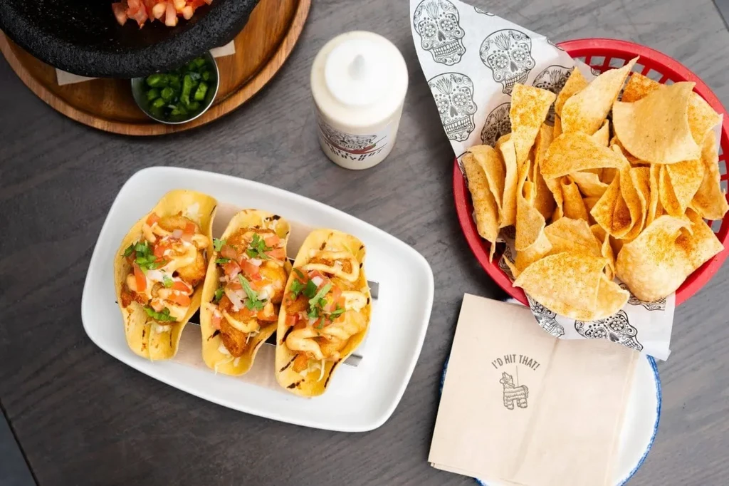 Overhead: Three fish tacos, salsa, chips, and peppers on a table.