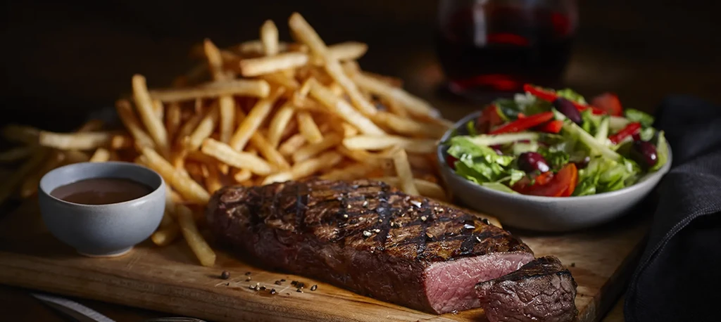 Medium-rare grilled steak, fries, salad, and sauce on a board.