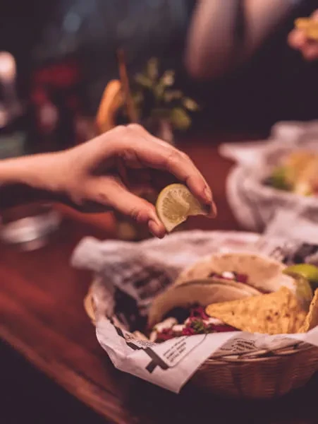 Hand squeezing lime wedge over tacos in a rustic basket.