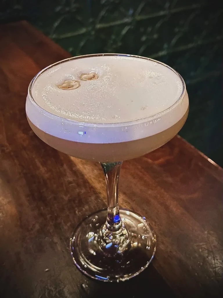 Amber cocktail with white foam and two garnishes in a coupe glass.
