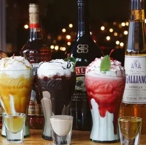 Three colorful dessert cocktails with whipped cream, shot glasses, and liquor bottles.