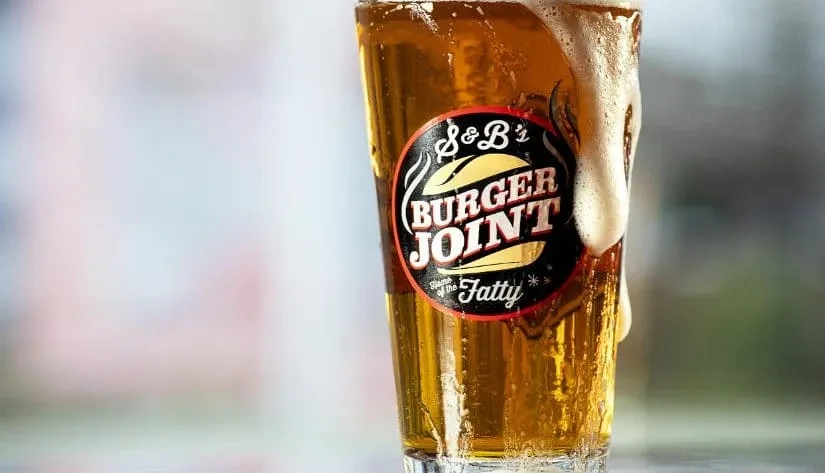 Pint glass of golden beer, frothy head overflowing, S&B's Burger Joint label.