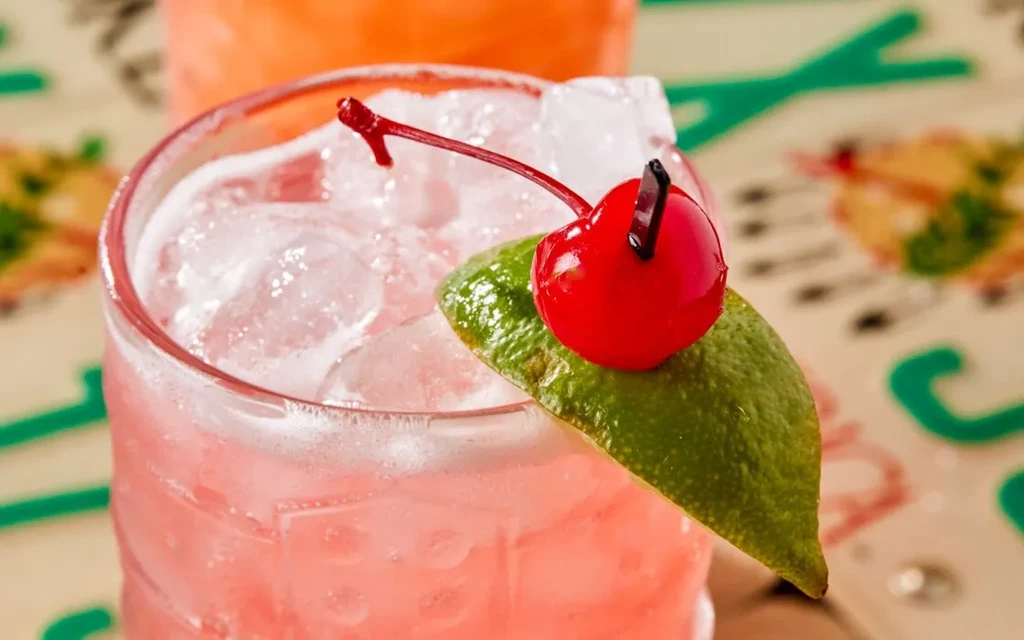 Close-up pink cocktail with cherry, lime, and ice in textured glass.