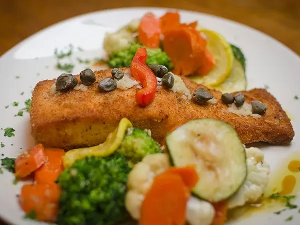 Plated breaded fish, capers, red pepper, and colorful steamed vegetables.