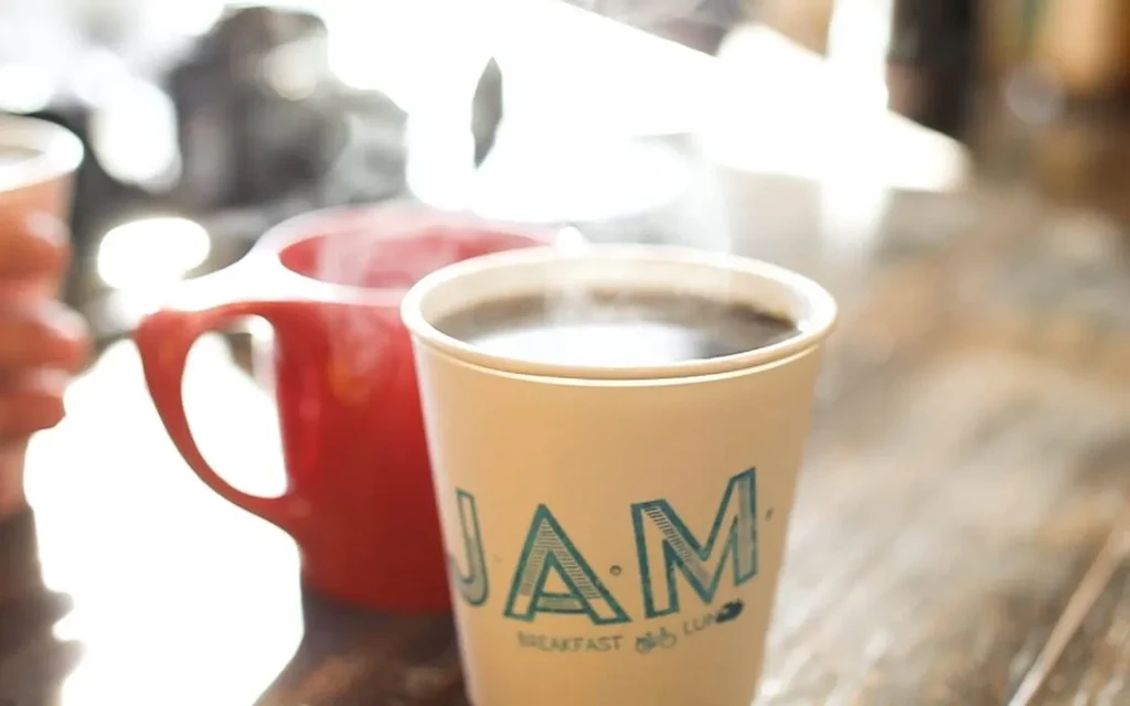 Steaming coffee in a "JAM BREAKFAST & LUNCH" paper cup on wood.