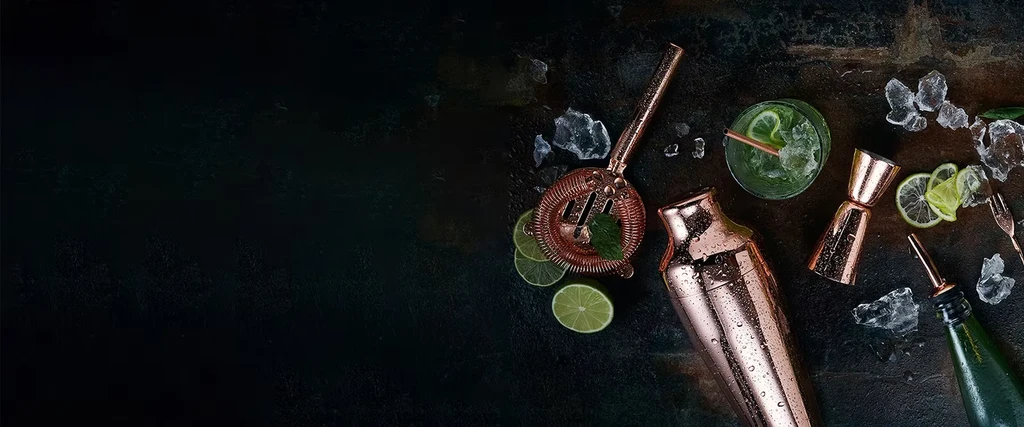 Top-down view of a cocktail, lime, mint, and copper bar tools.